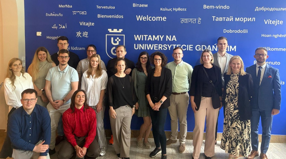 Na zdjęciu uwieczniono grupę pracowników i studentów zgromadzonych przed charakterystyczną ścianą powitalną Uniwersytetu Gdańskiego. Napis „Witamy na Uniwersytecie Gdańskim” otoczony jest tłumaczeniami słowa „witamy” w różnych językach, co podkreśla wielokulturowy i otwarty charakter uczelni. Osoby pozujące do zdjęcia reprezentują różnorodność – zarówno pod względem wieku, jak i stylu ubioru – co świadczy o integracyjnym i przyjaznym środowisku akademickim.  Lokalizacja: Uniwersytet Gdański, ściana powitalna z napisem „Witamy” w wielu językach.  Osoby: Grupa około 17 osób – zarówno kobiety, jak i mężczyźni – ustawionych w dwóch rzędach.  Styl ubioru: Elegancki i casualowy – koszule, marynarki, sukienki, spodnie w kant, ale również luźniejsze stroje, jak swetry czy koszulki polo.  Atmosfera: Profesjonalna, ale przyjazna – osoby pozują z uśmiechem, co nadaje zdjęciu ciepły ton.  Przeznaczenie zdjęcia: na potrzeby dokumentacji wydarzenia.
